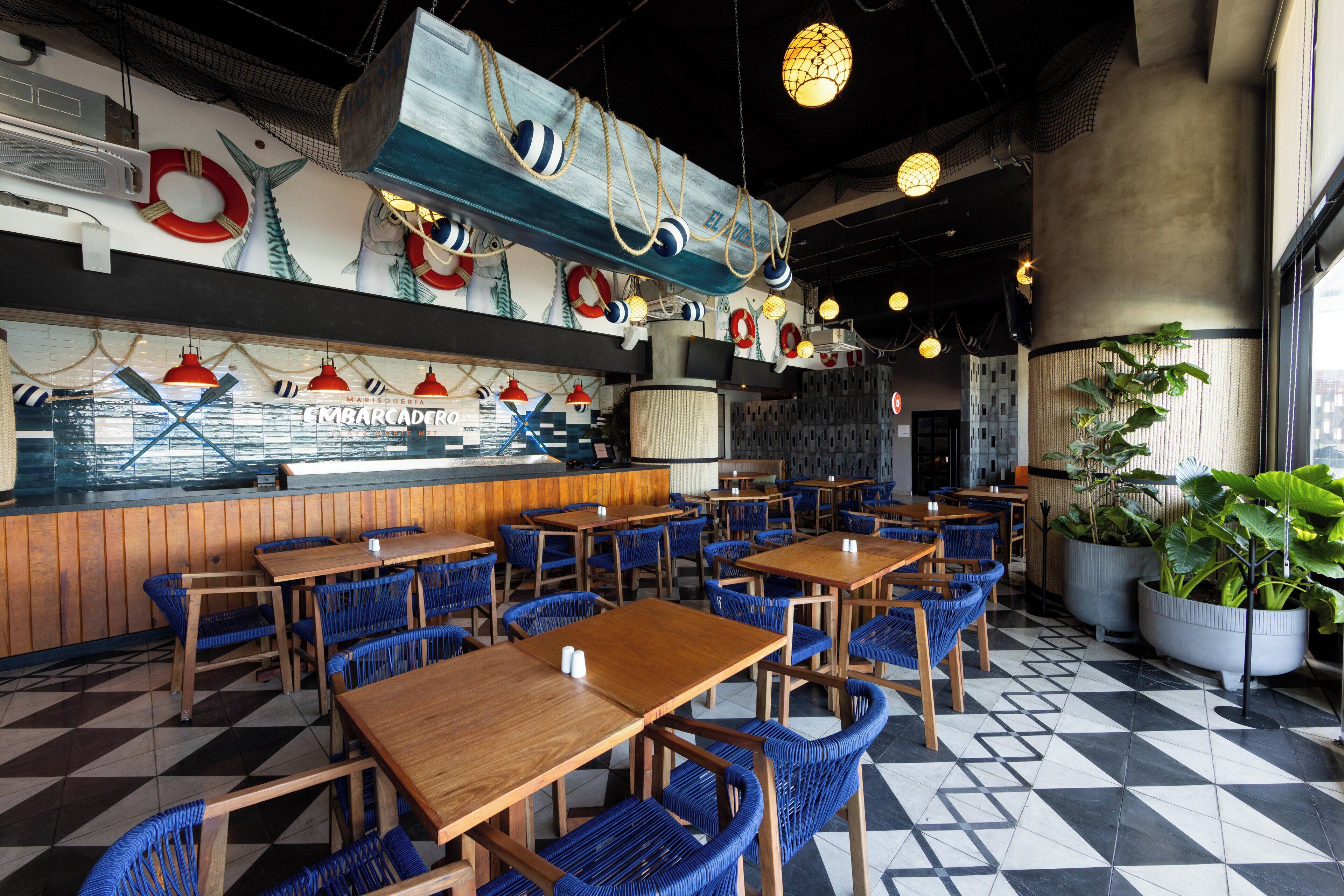En restaurant med træborde og blå stole, maritim indretning og et skakternet gulv.