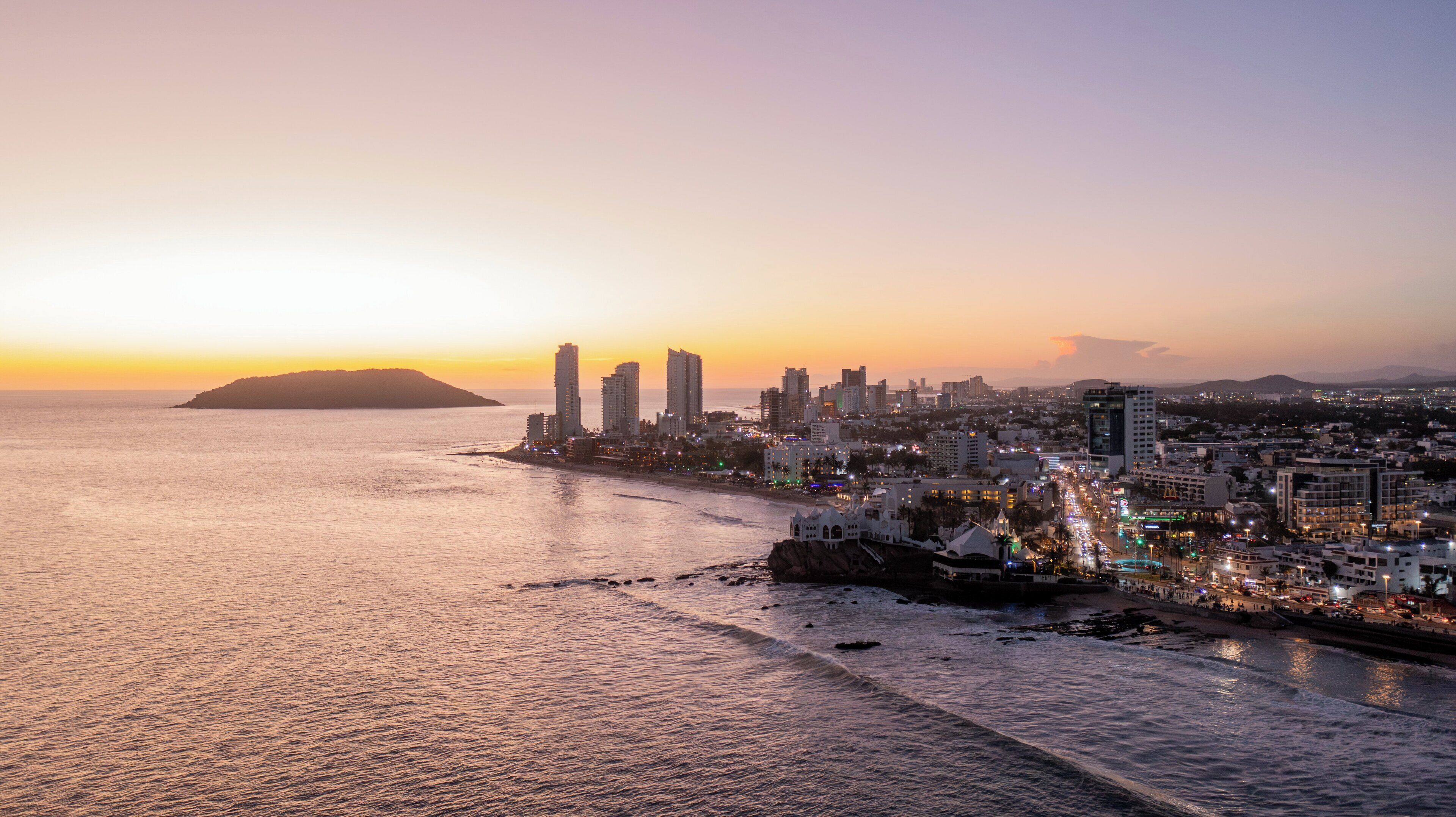 A coastal cityscape at sunset with high-rise buildings, a beach, and a mountain in the distance.