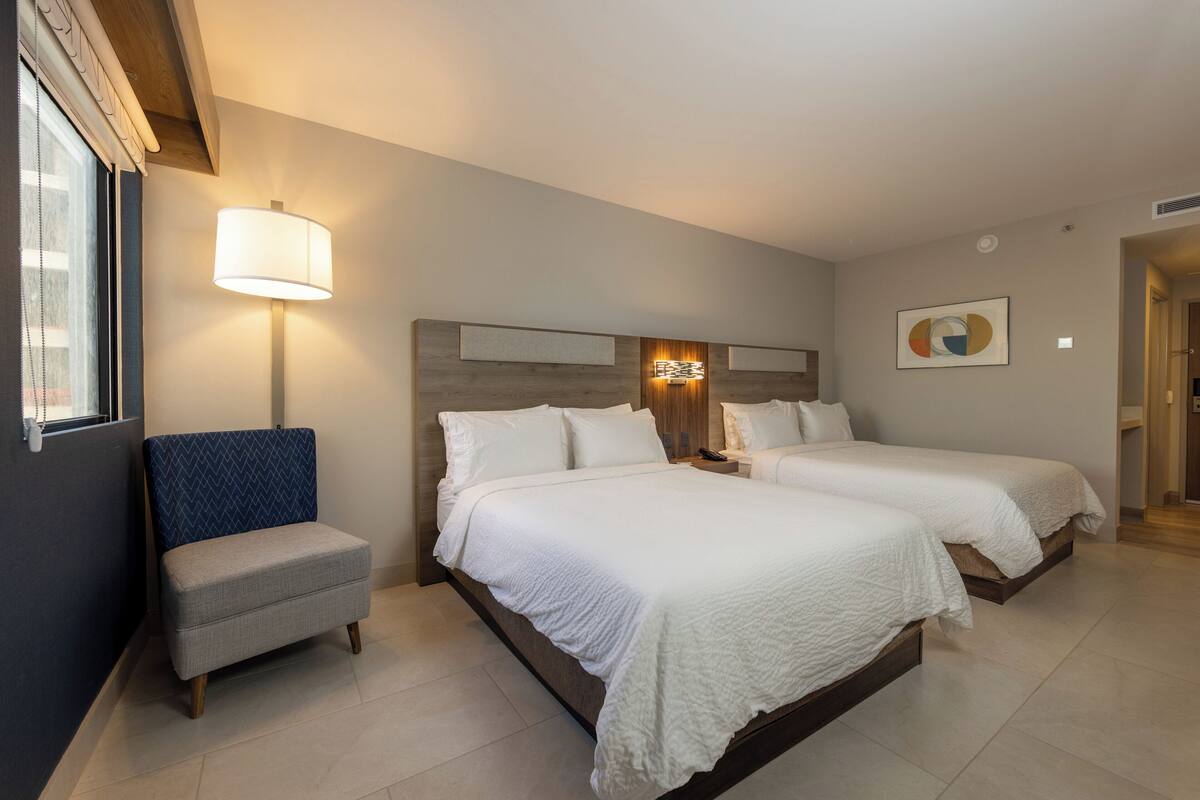 Ett hotellrum med två sängar, ett sängbord, en lampa, en stol och ett konstverk som hänger på väggen.