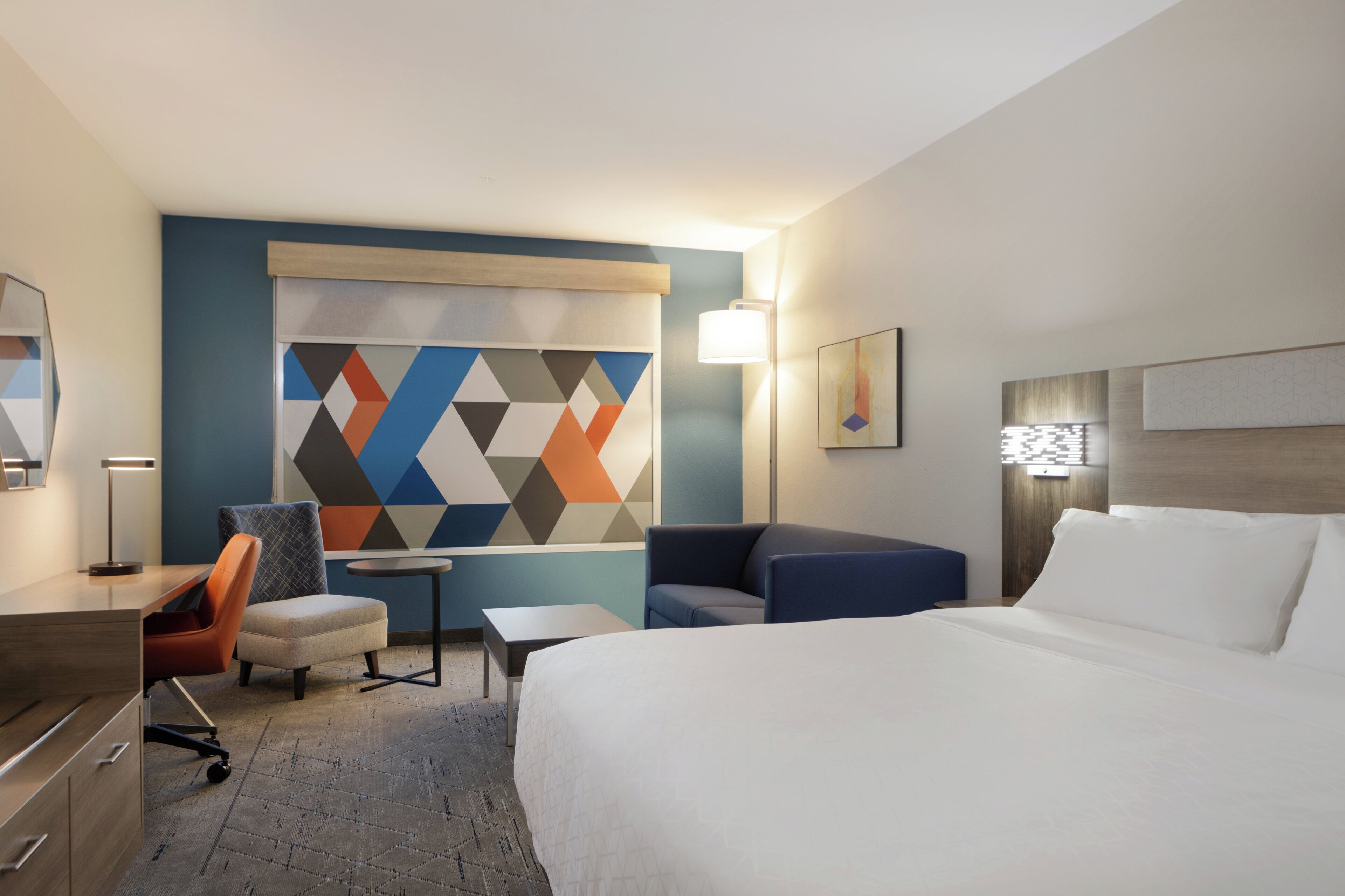 Ein Hotelzimmer mit einem Bett, einem Schreibtisch, einem Sessel, einem kleinen Tisch und einem farbenfrohen Wandbild mit geometrischen Formen.
