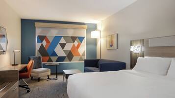 Ein Hotelzimmer mit einem Bett, einem Schreibtisch, einem Sessel, einem kleinen Tisch und einem farbenfrohen Wandbild mit geometrischen Formen.