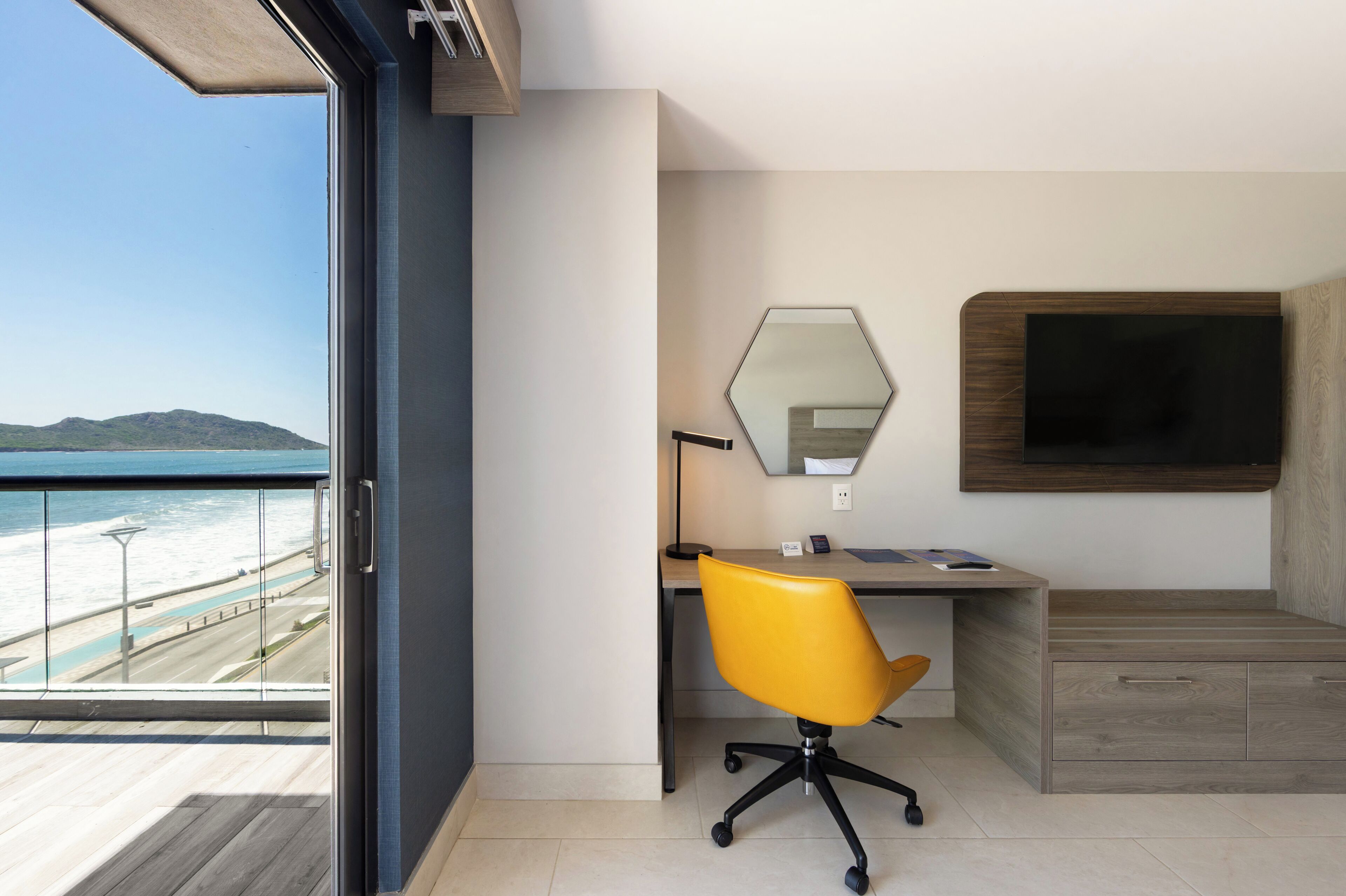 A modern room with a desk, chair, and TV, overlooking a beach.
