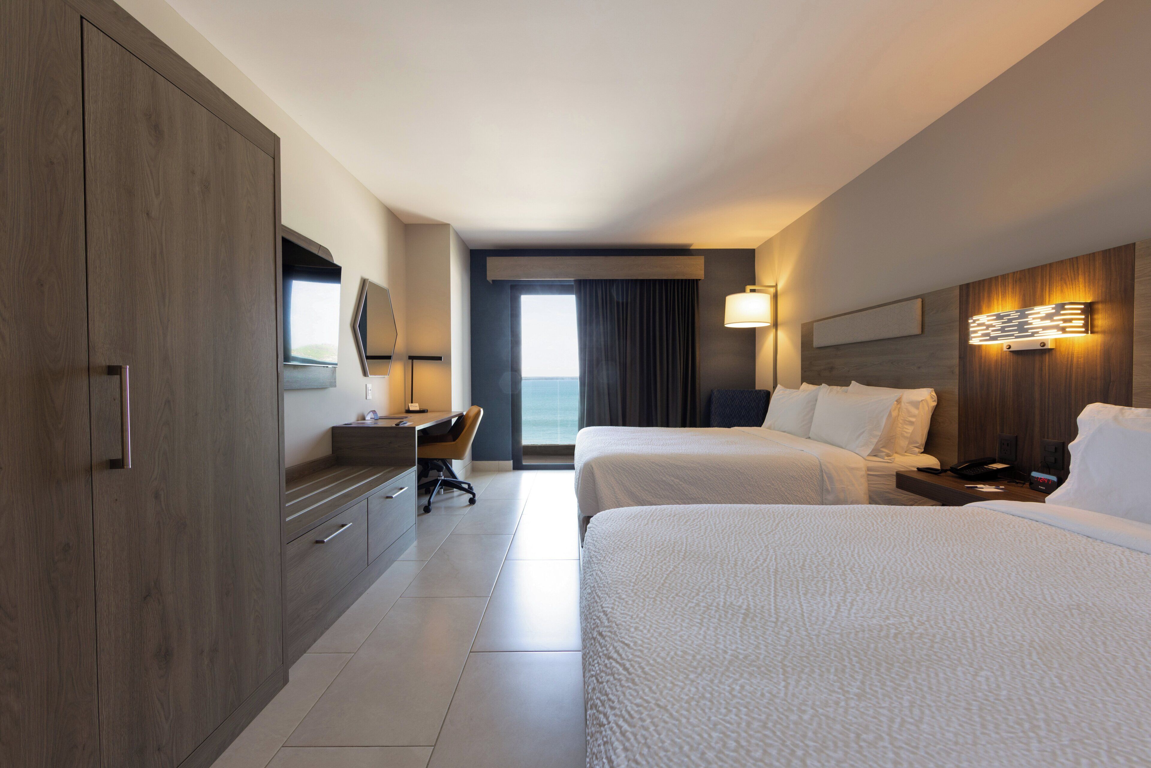 A hotel room with two beds, a desk, a chair, a large wardrobe, a TV, and a view of the outdoors.