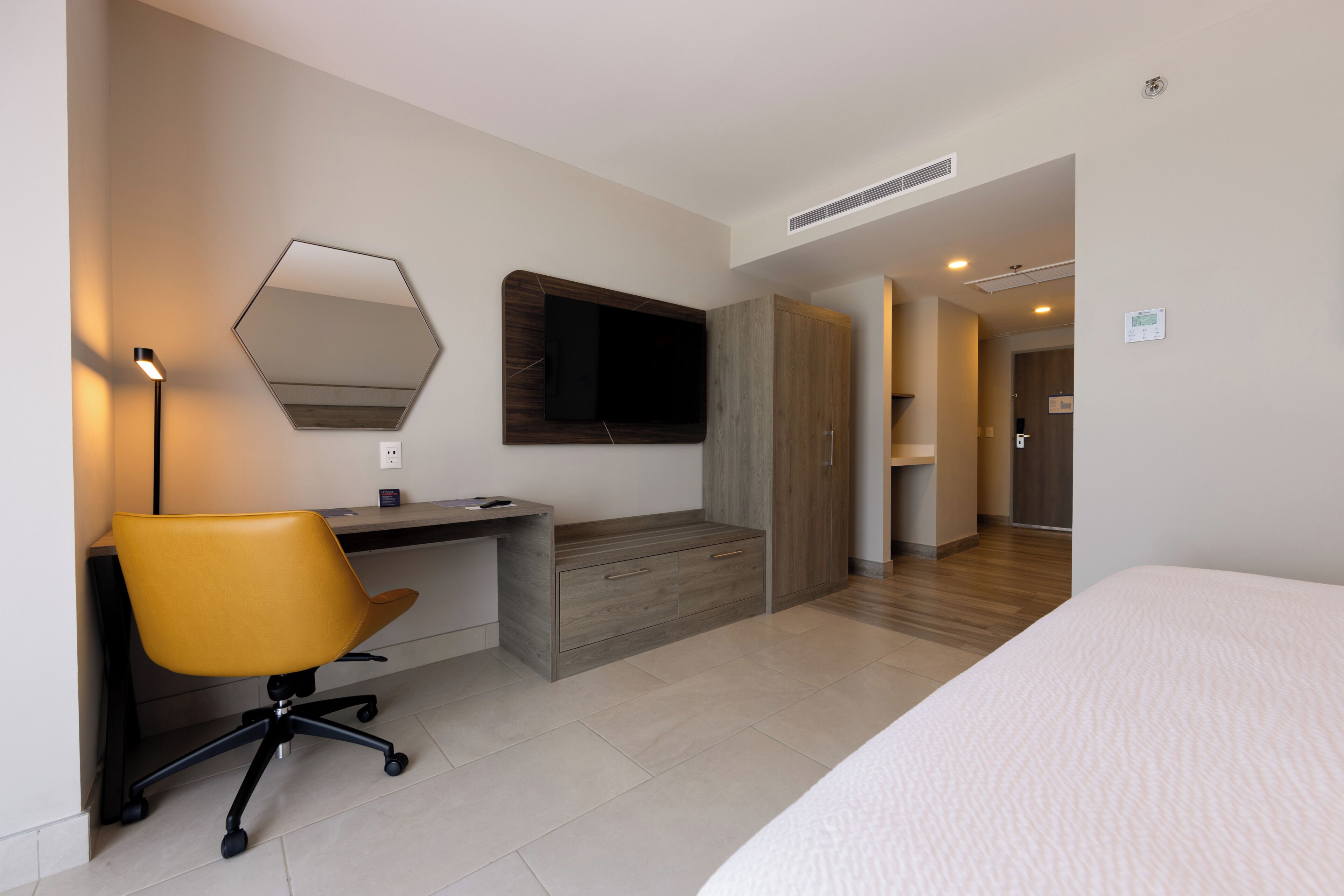 A modern hotel room with a bed, a desk with a chair, a large mirror, and a wardrobe.