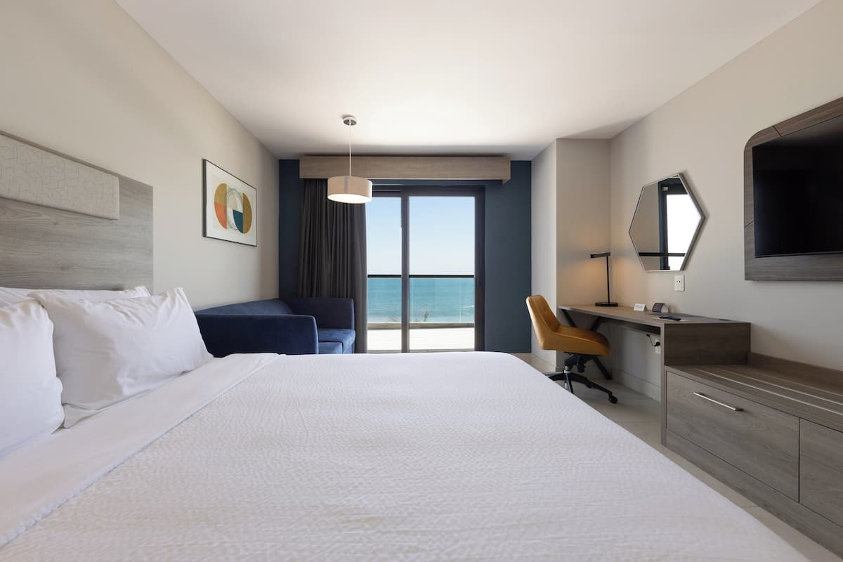 Ett hotellrum med en stor säng, ett skrivbord, en stol och utsikt över havet.