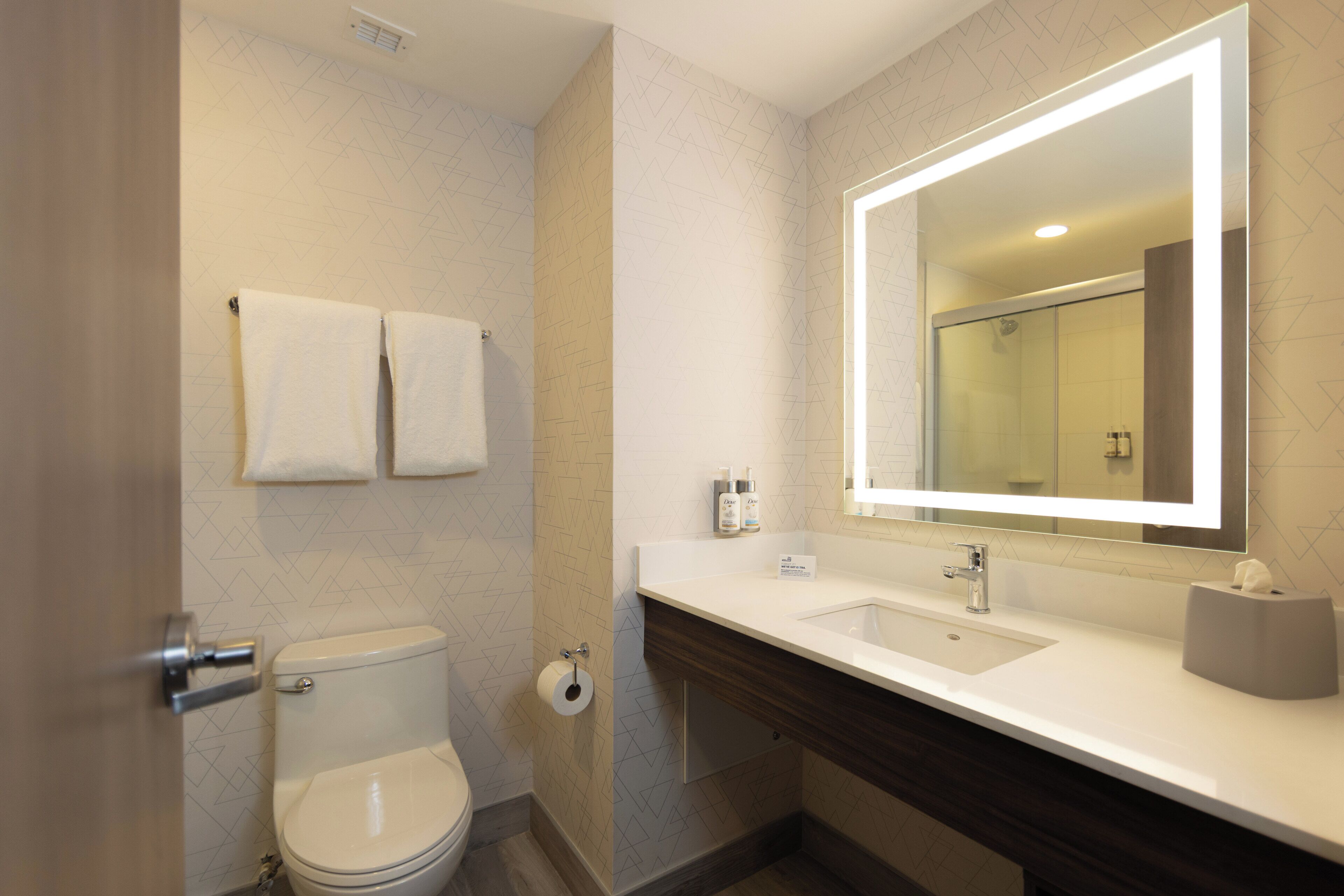 Un baño moderno con inodoro, dos toallas, lavamanos, espejo y ducha.
