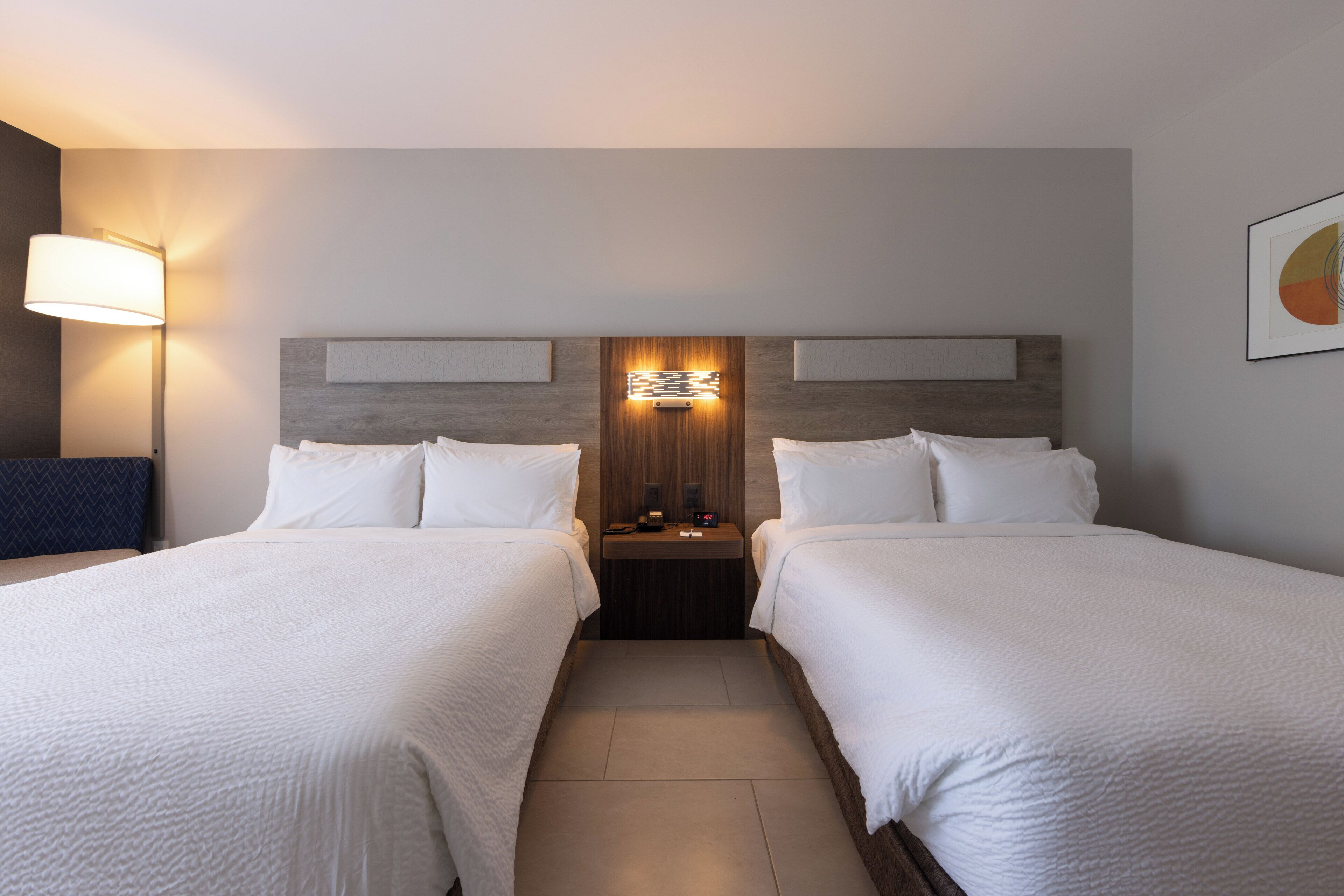 A hotel room with two beds, a nightstand, a wall-mounted light, and a framed picture.