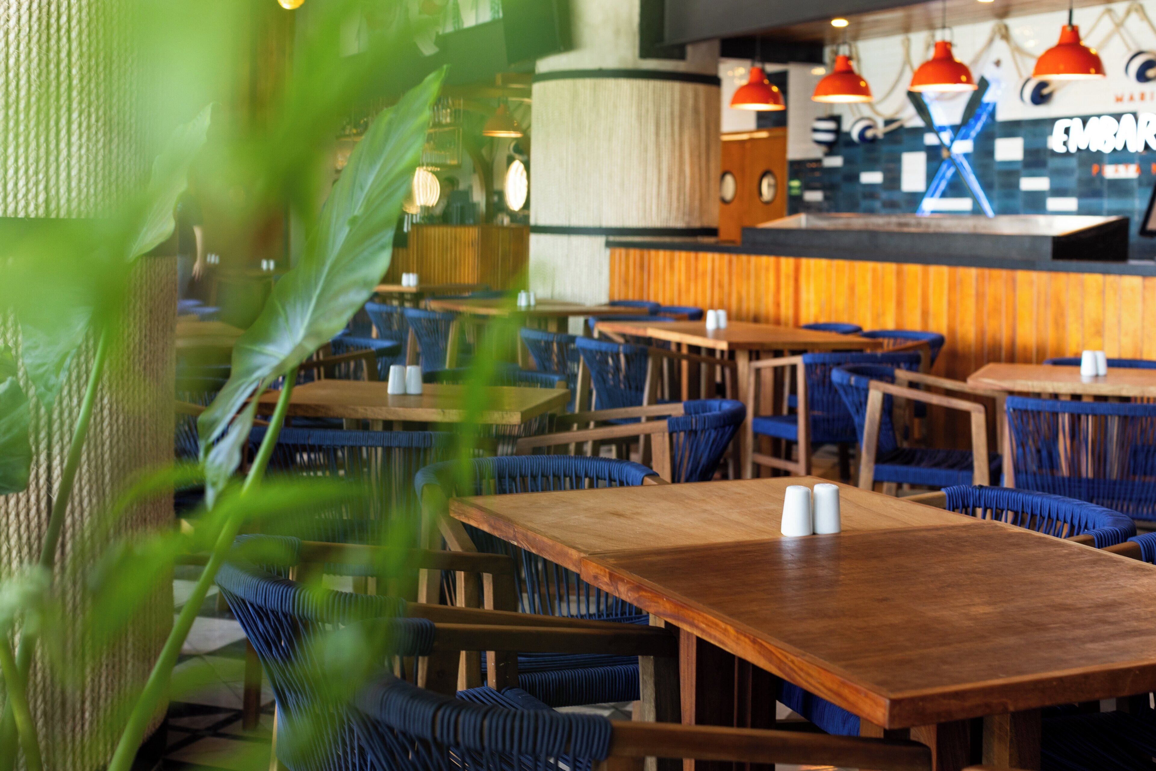 A restaurant interior with wooden tables and blue upholstered chairs, a bar area, and hanging red pendant lights.