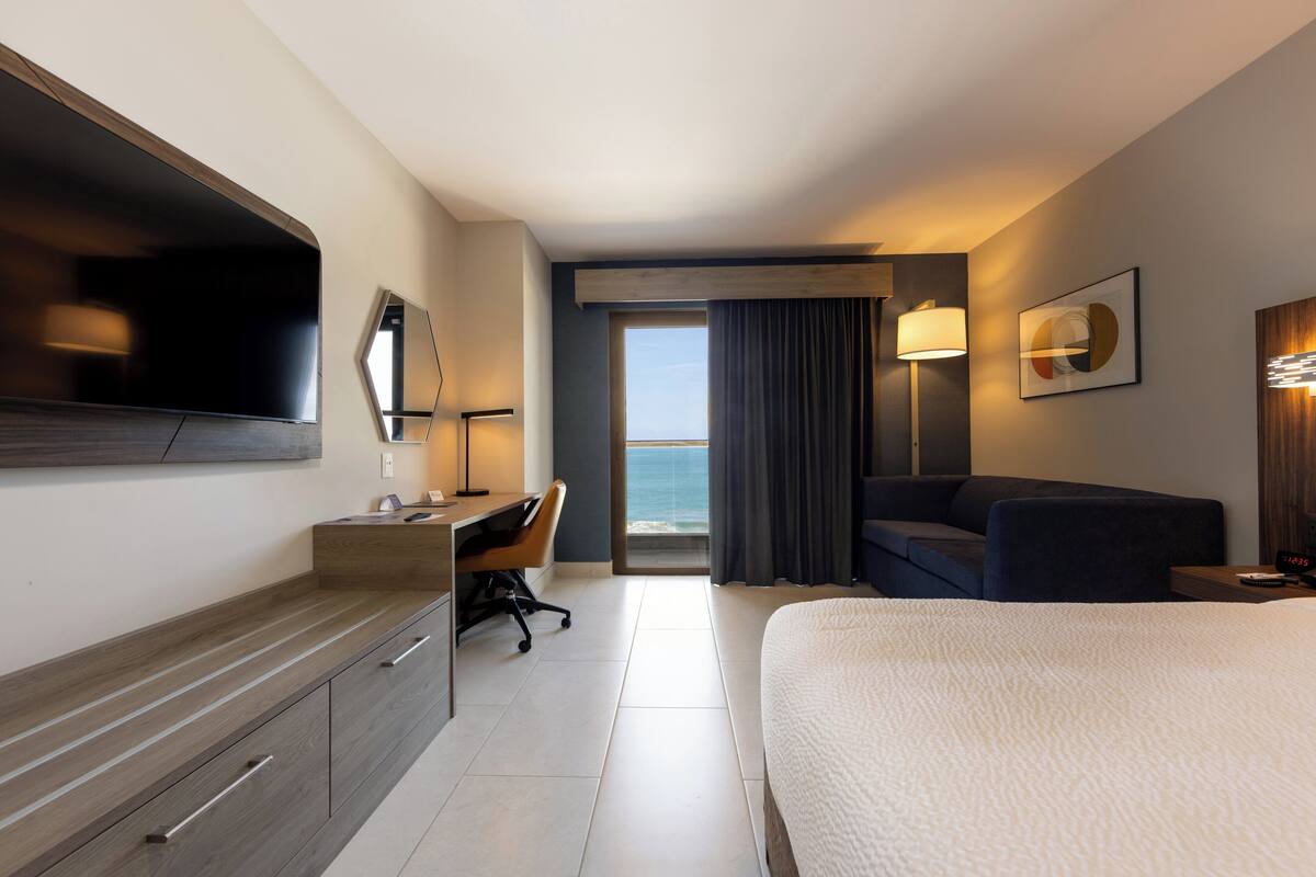 Ett hotellrum med en säng, ett skrivbord, en stol och en stor TV.