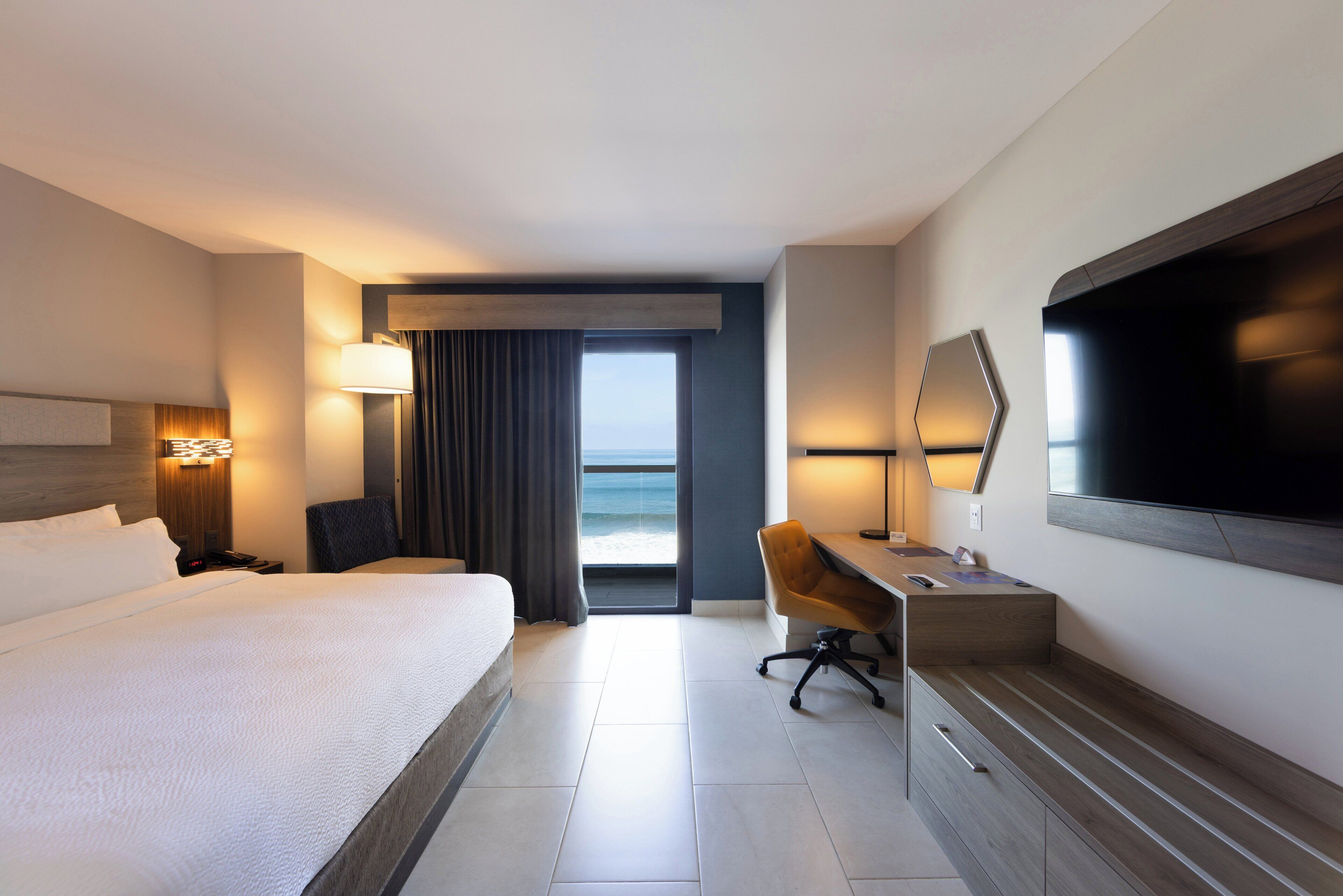 A hotel room with a bed, desk, chair, TV, and a view of the ocean.