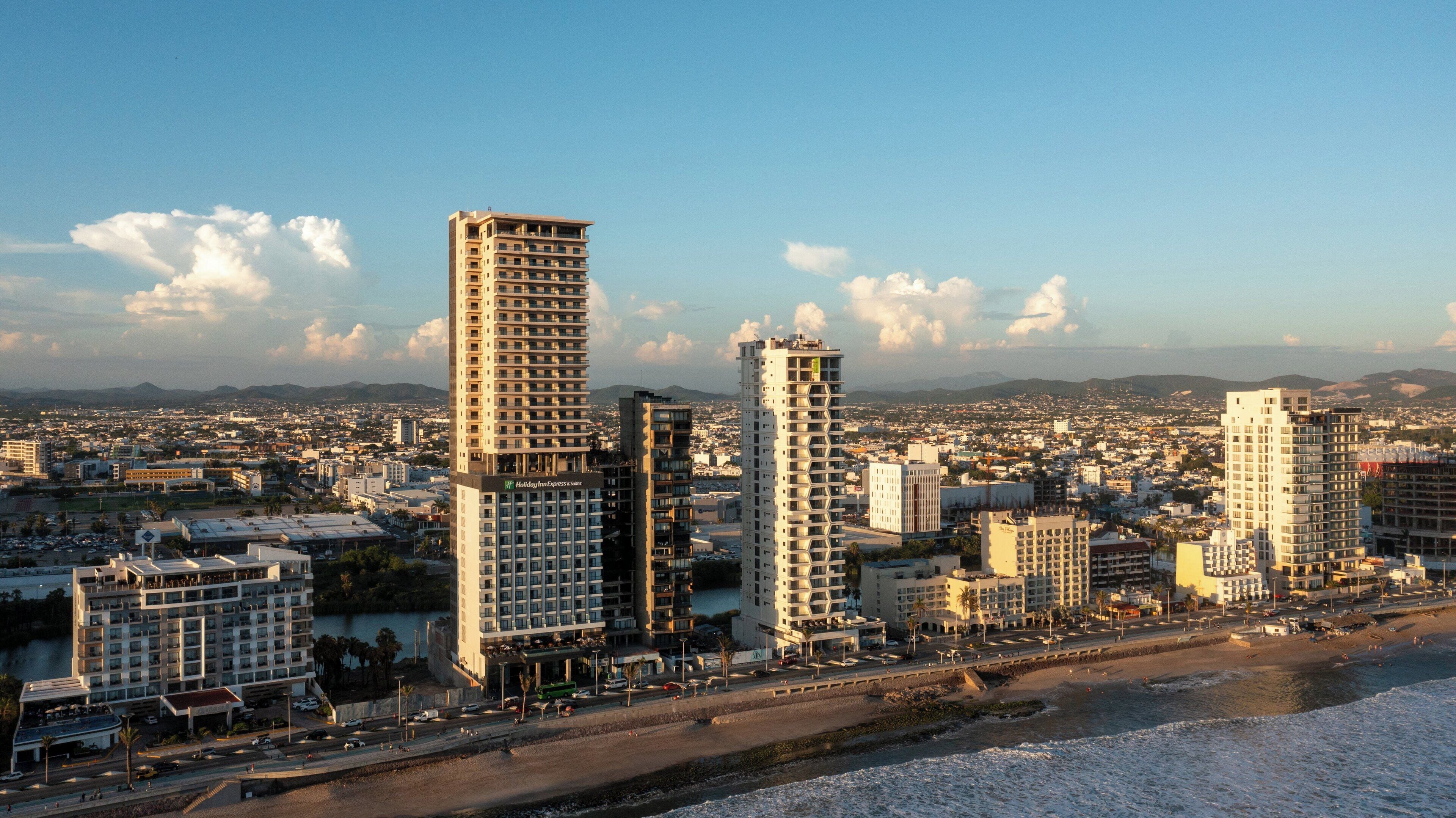 A high-rise hotel with multiple floors, a prominent Holiday Inn Express & Suites sign, and a view of the cityscape and beach in the background.