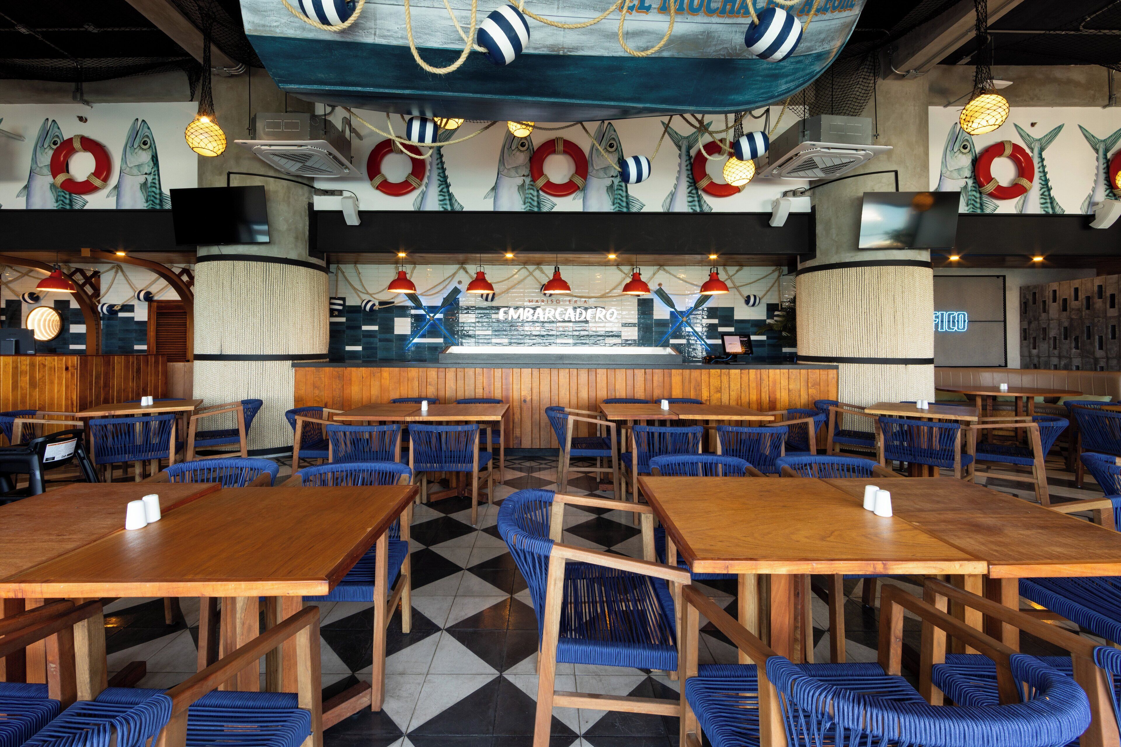Un restaurant avec des tables en bois et des chaises bleues, un comptoir illuminé par un néon, et des poissons décoratifs et des bouées accrochées aux murs.