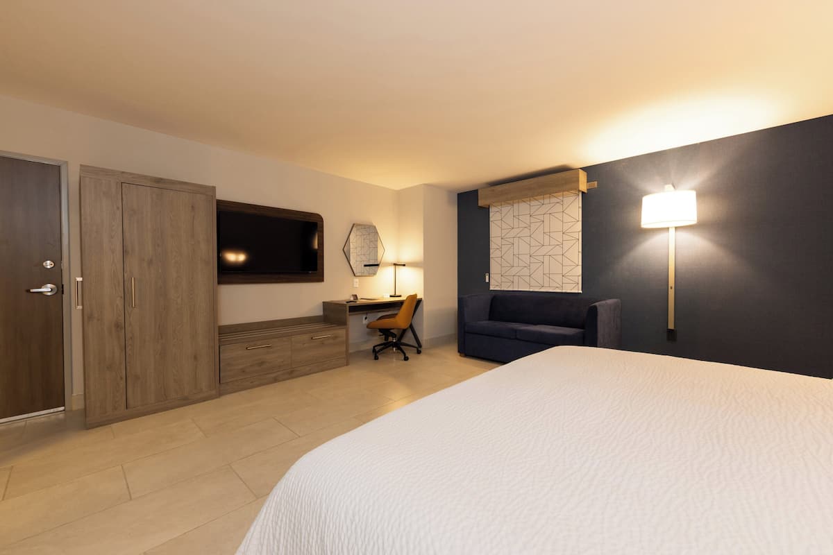 Ett hotellrum med en säng, skrivbord, stol, garderob, TV och en väggmonterad lampa.