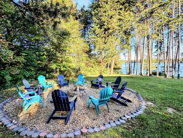 A circular seating arrangement with chairs around a fire pit, surrounded by a gravel path and a grassy area, with trees and a body of water in the background.