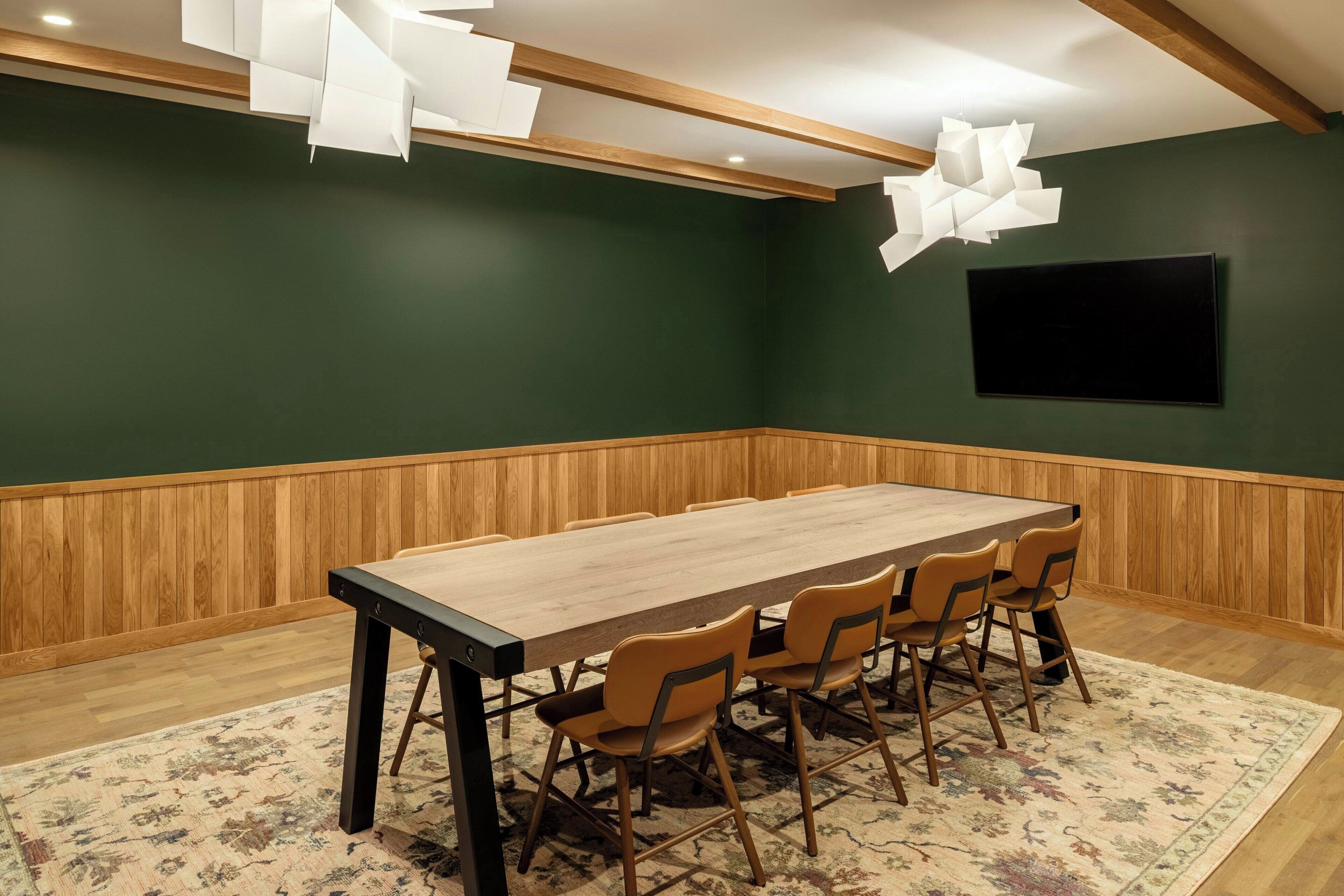 A conference room with a long table, six chairs, a wooden floor, a green wall, and a mounted TV.