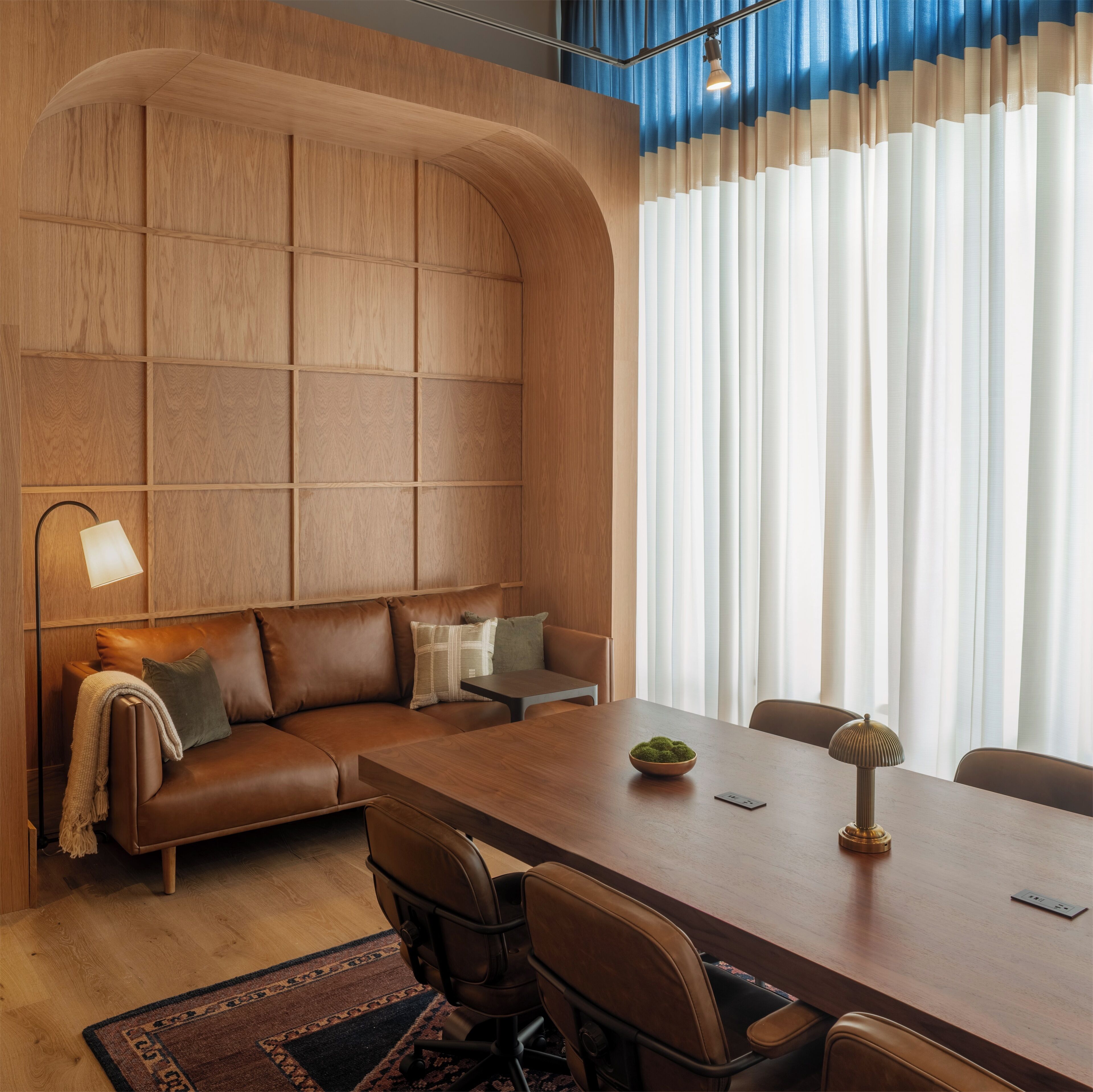A modern interior with a wooden panel wall, a brown leather sofa, a wooden dining table with chairs, and a large window with white curtains.