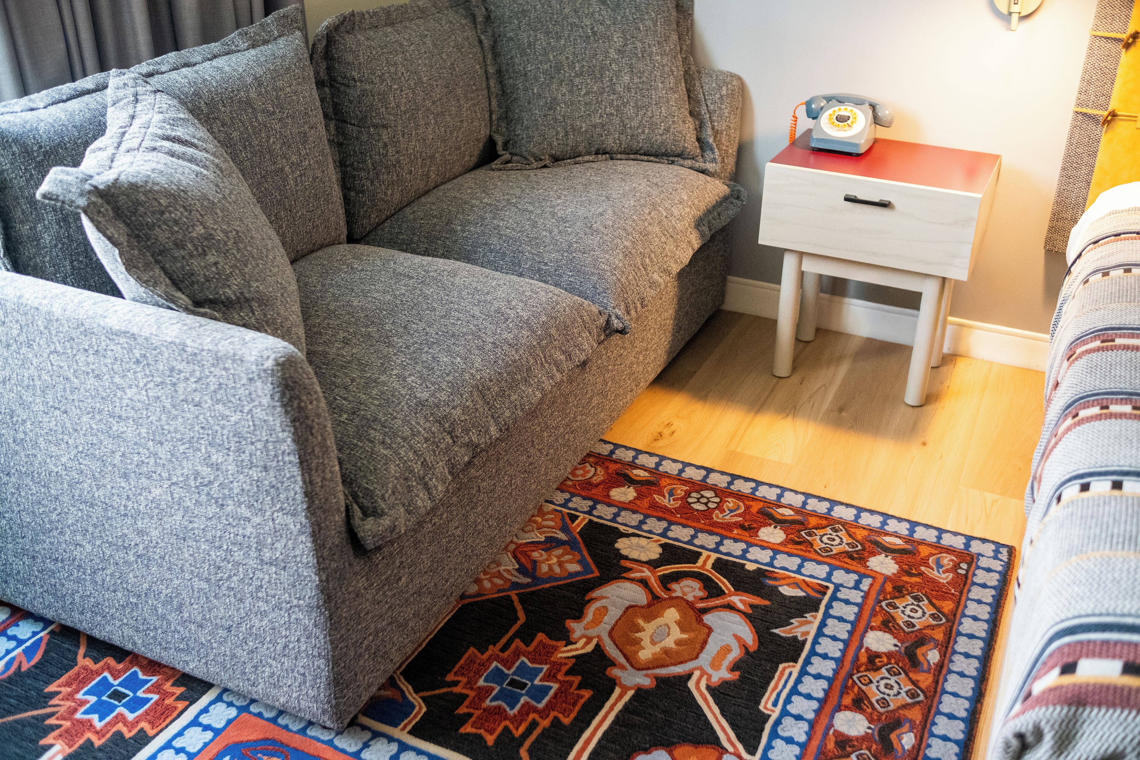 A cozy living room with a grey sofa, a small wooden side table with a telephone, and a patterned rug.