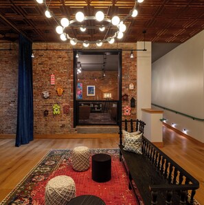 A cozy room with a brick wall, a wooden ceiling, and a staircase. There are hanging lights, a bench, and a patterned rug.