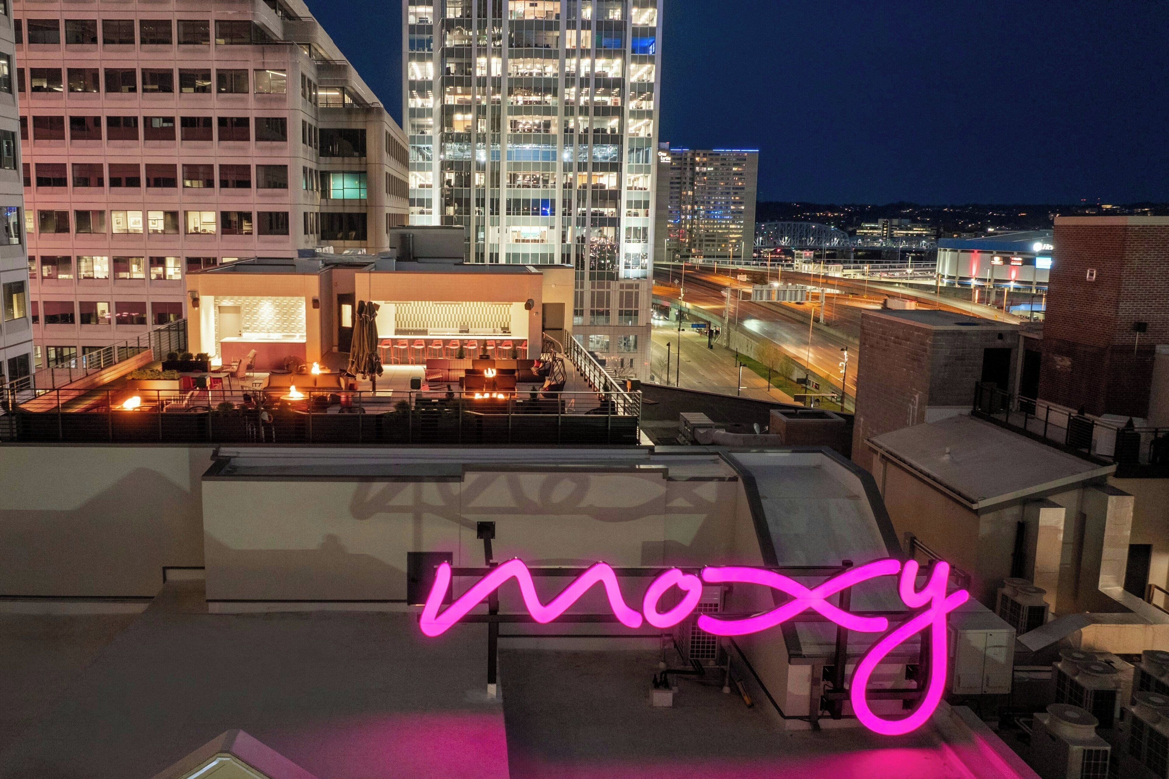 A rooftop bar with a neon sign, modern buildings, and a cityscape in the background.
