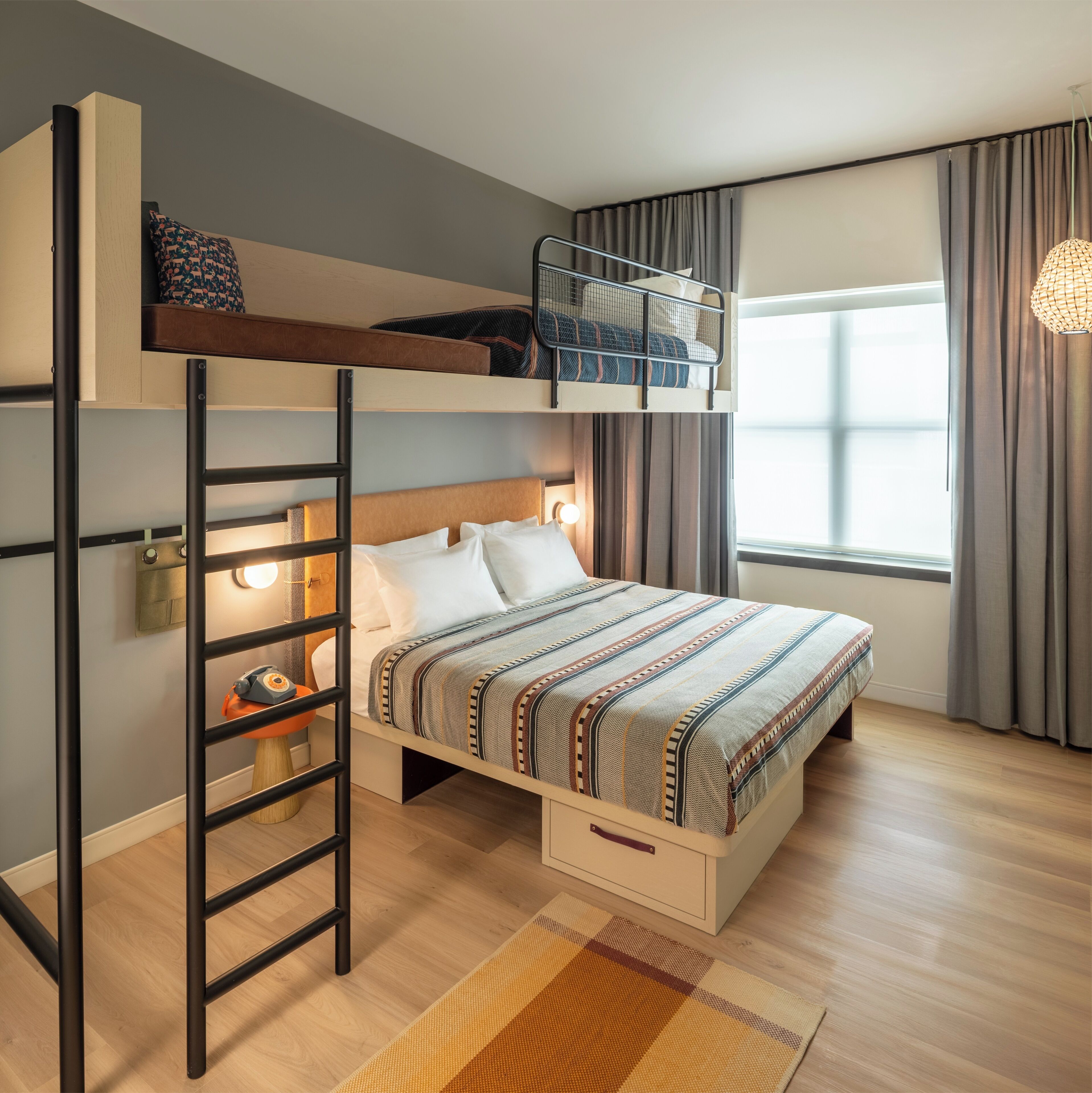 A bunk bed room with a ladder, a bedside table, a lamp, and a window with curtains.