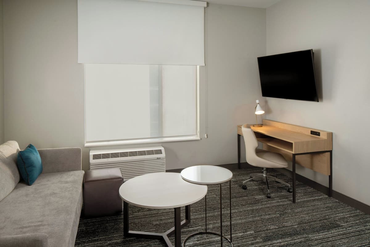 A hotel room with a sofa, two small tables, a desk with a chair, a TV mounted on the wall, and a window with blinds.