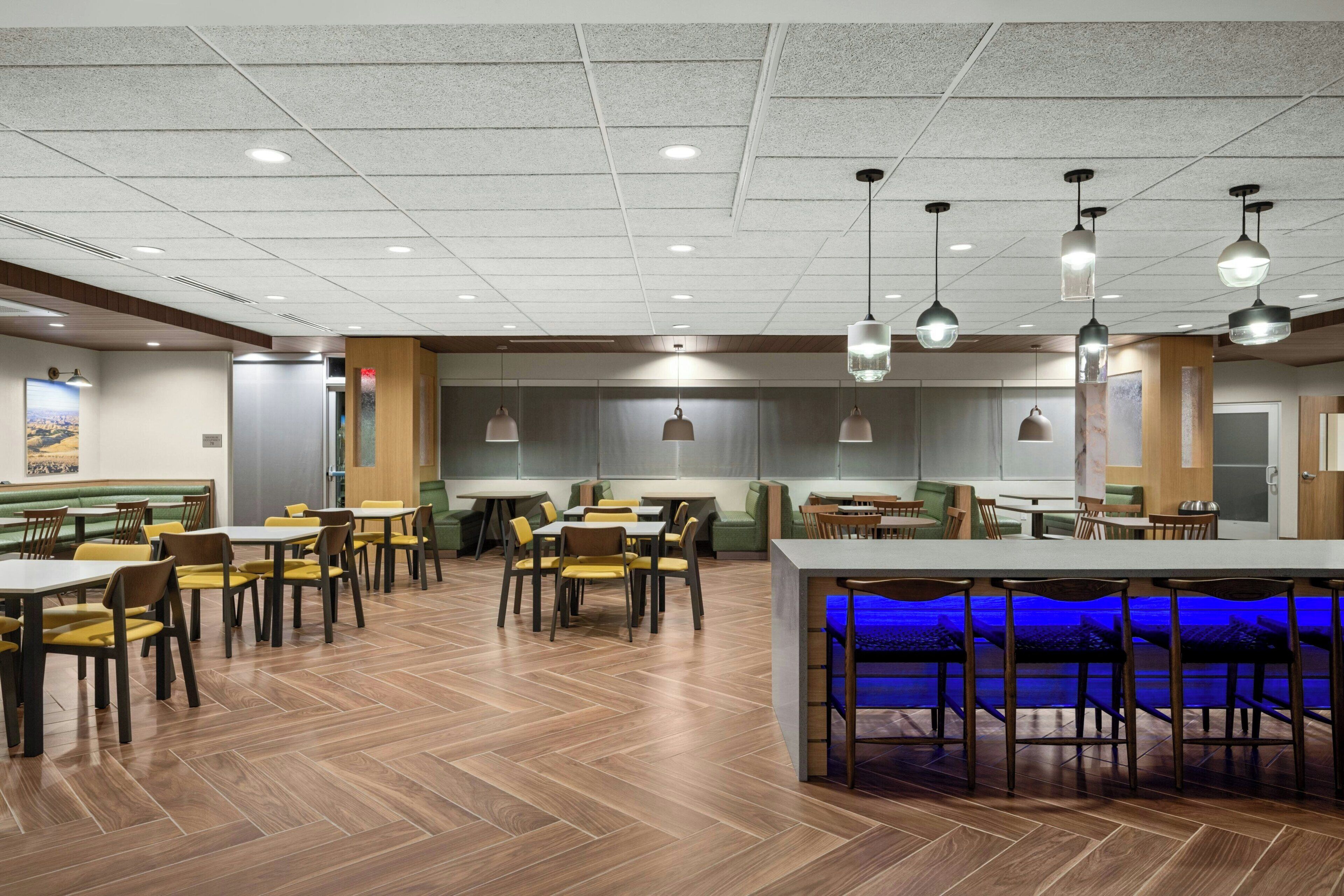 A spacious dining area with multiple tables and chairs, recessed ceiling lights, and a blue-lit bar area.