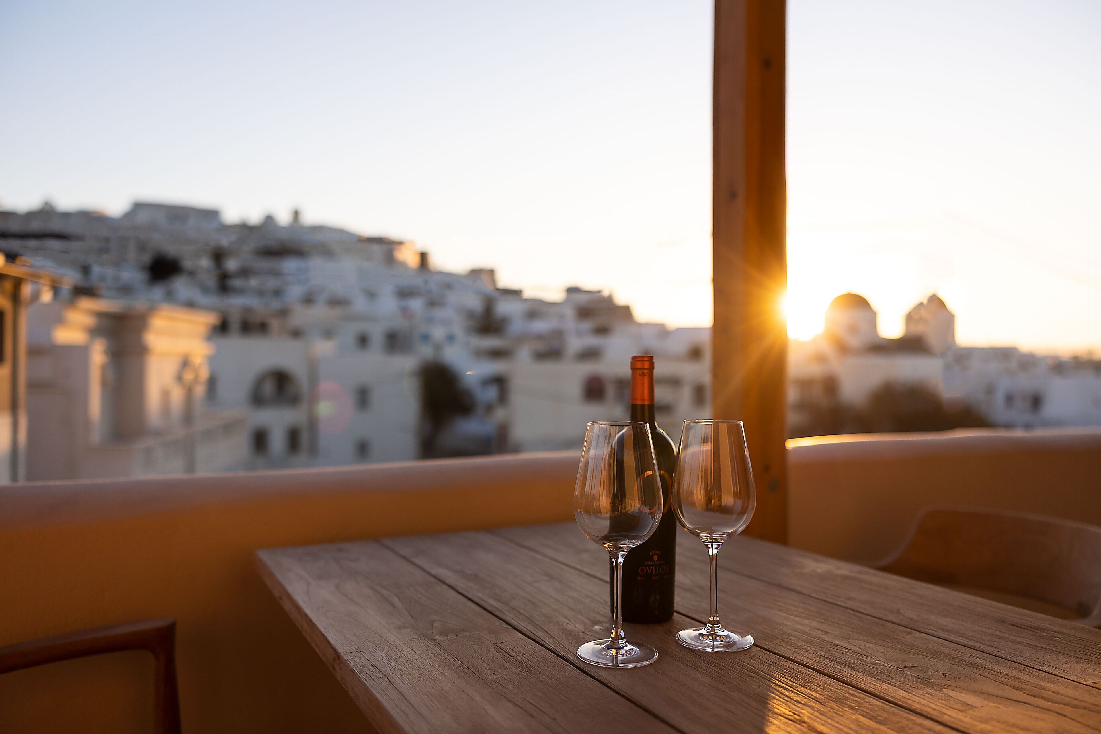 Ein Holztisch mit einer Flasche und zwei Gläsern auf einem Balkon mit Blick auf eine Stadtlandschaft bei Sonnenuntergang.