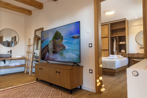A modern room with a large flat-screen TV, wooden furniture, a bed, and a bathroom with a wooden cabinet and shelves.