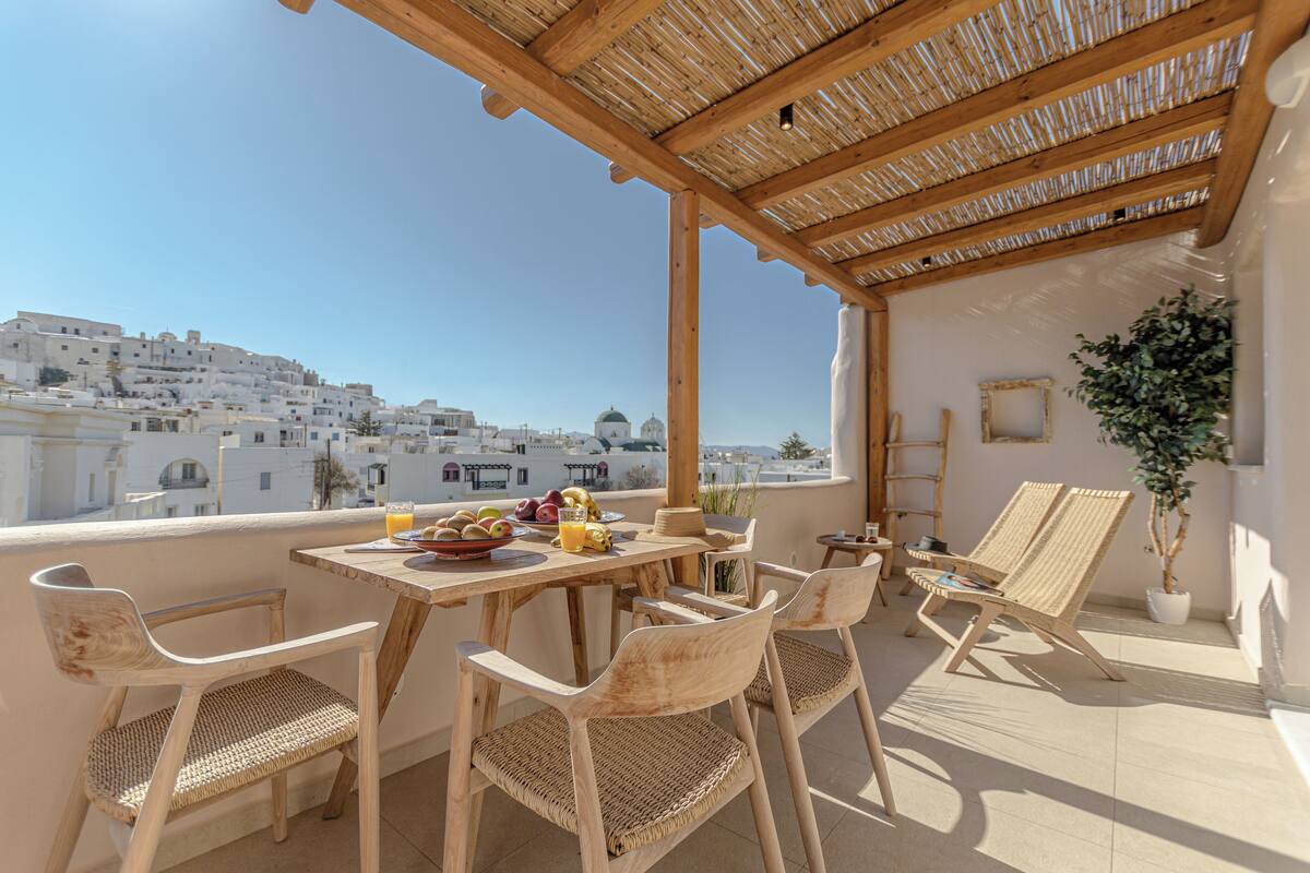 A terrace with wicker furniture, a table with fruits and drinks, and a view of a white town.