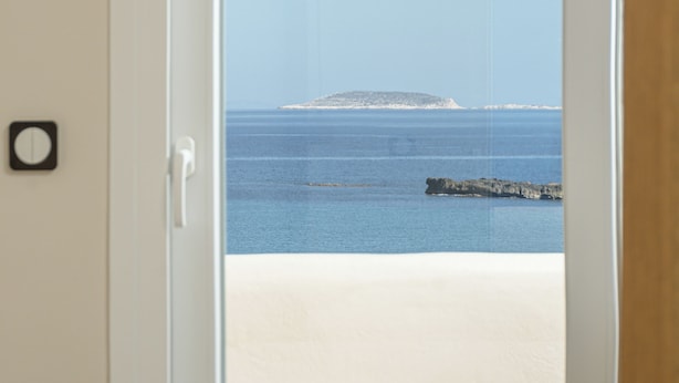 A room with a bed, a hat, and a view of the beach and sea through an open door. - Almi of Naxos (Naxos)