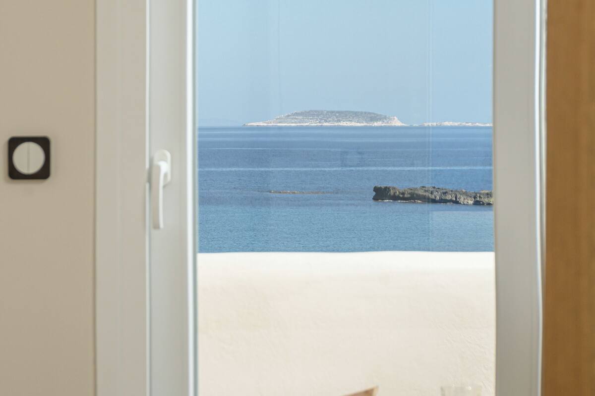 A room with a bed, a hat, and a view of the beach and sea through an open door.