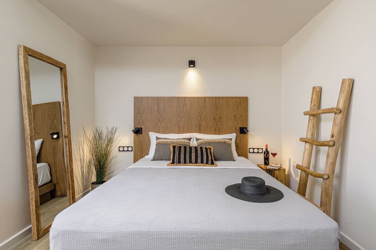 A neatly arranged bedroom with a large bed, a bedside table with a bottle and glass, a wooden ladder, and a mirror.