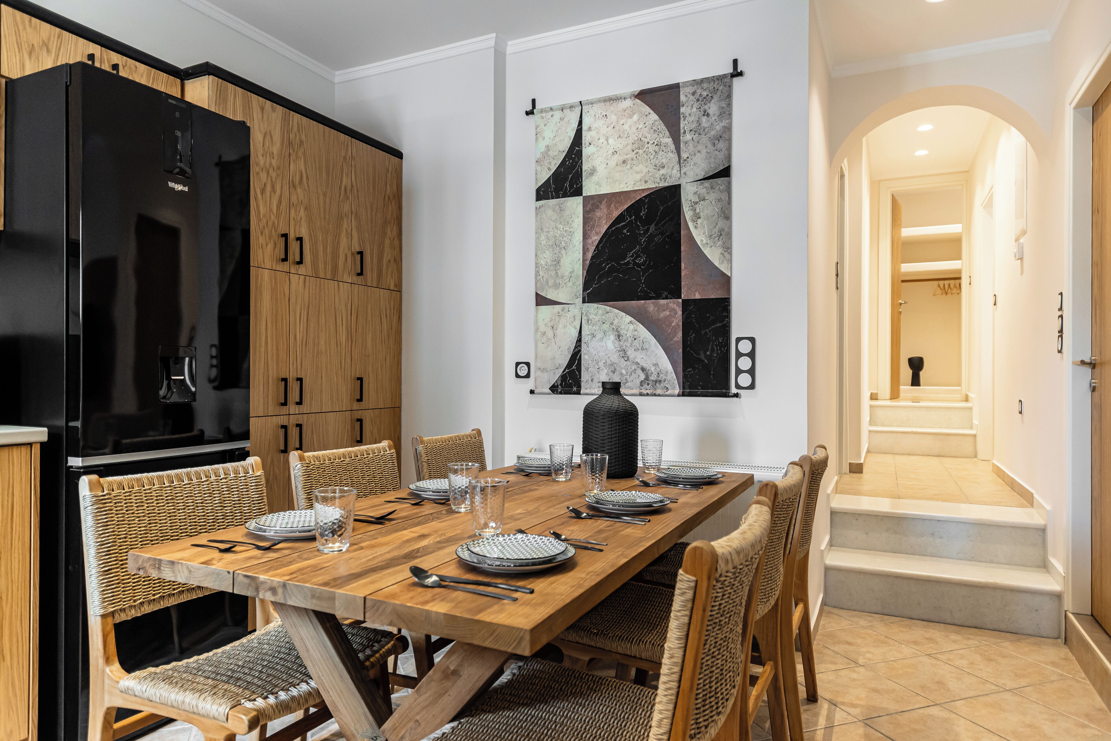 Un coin repas avec une table en bois dressée pour six personnes, entourée de chaises en osier. Un réfrigérateur noir est visible sur la gauche.