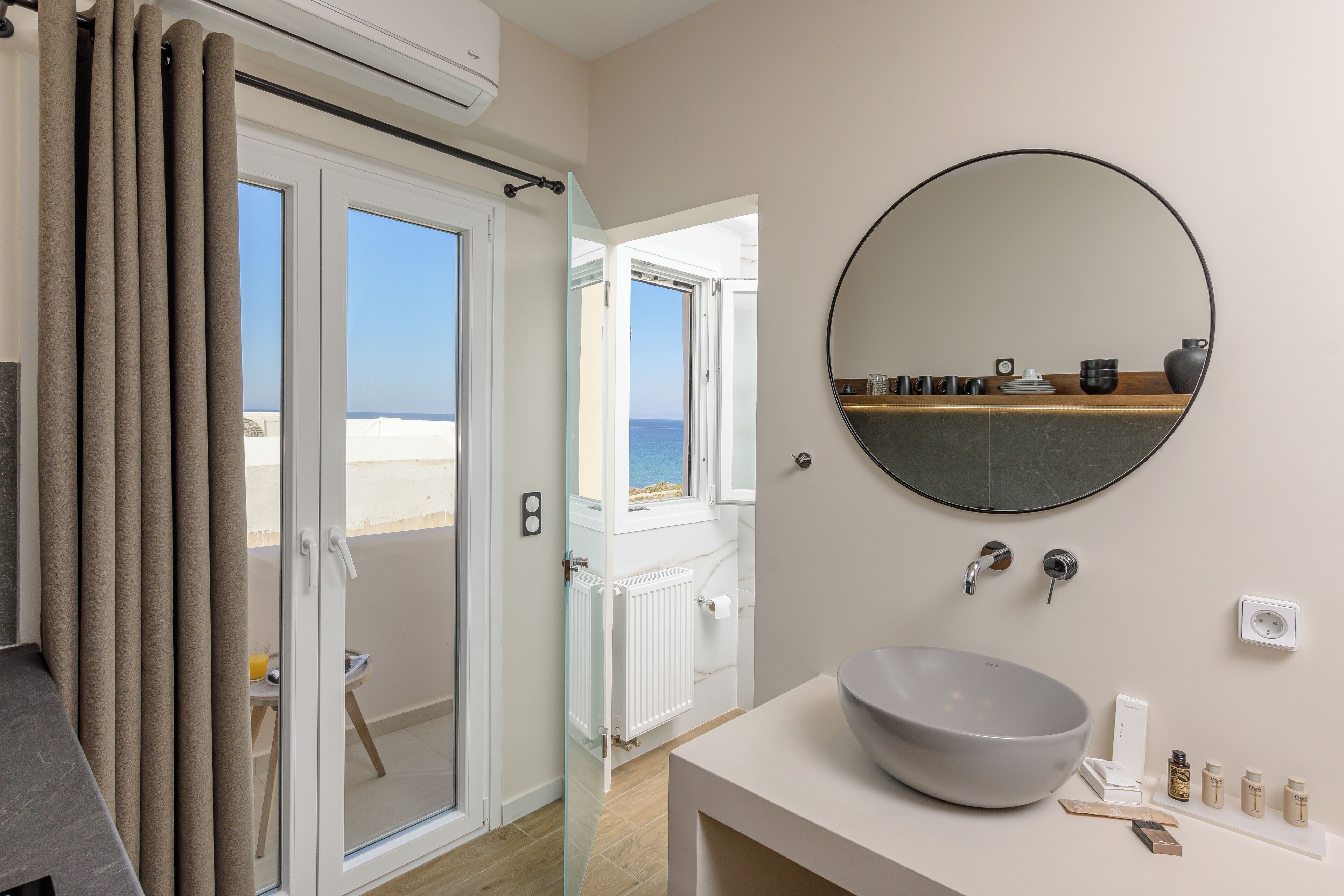 A bathroom with a large round mirror, a bowl sink, and a window overlooking the sea.