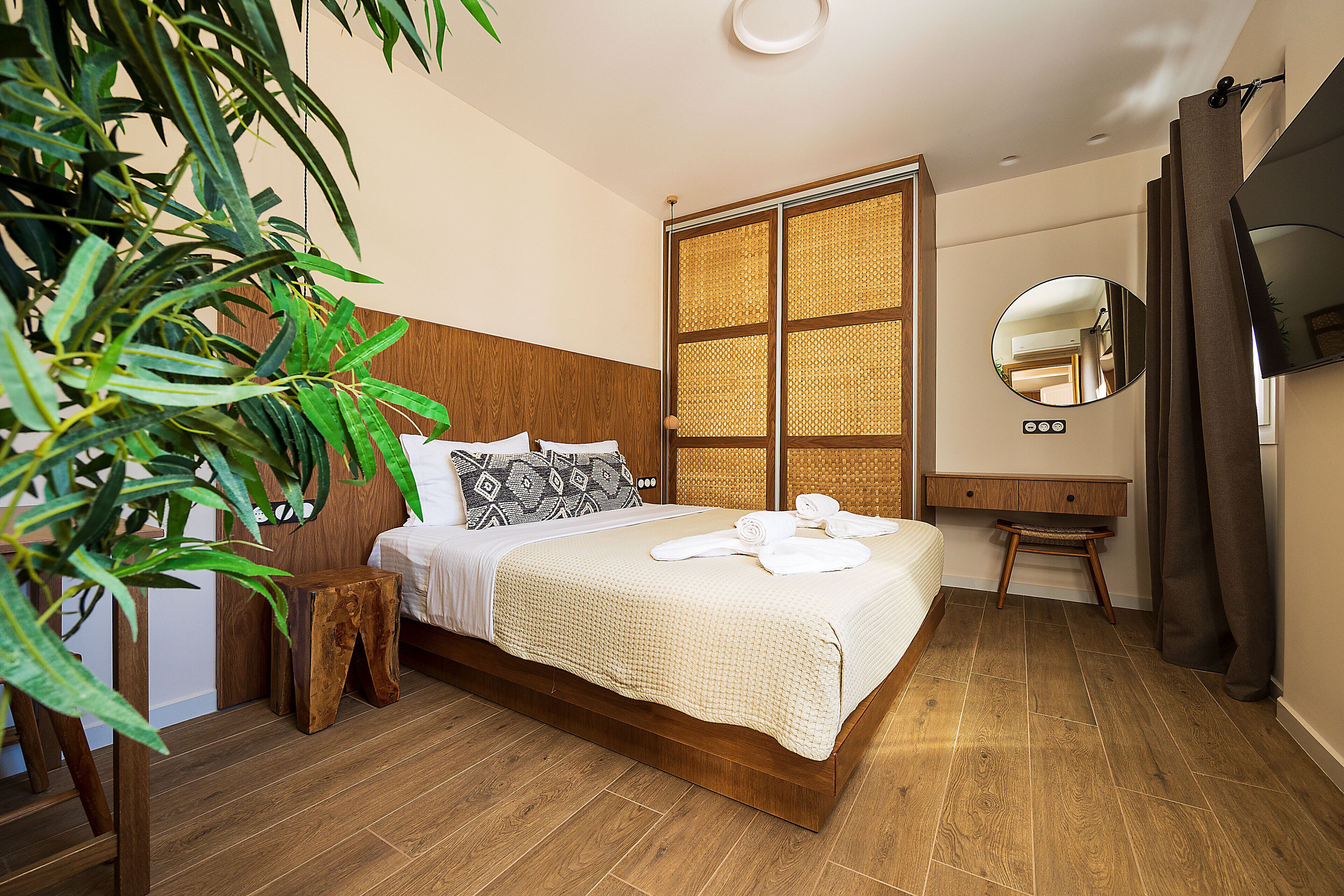 A neatly made bed with white linens and a rolled towel, a wooden nightstand, a large mirror, and a plant in a modern bedroom with wooden flooring.