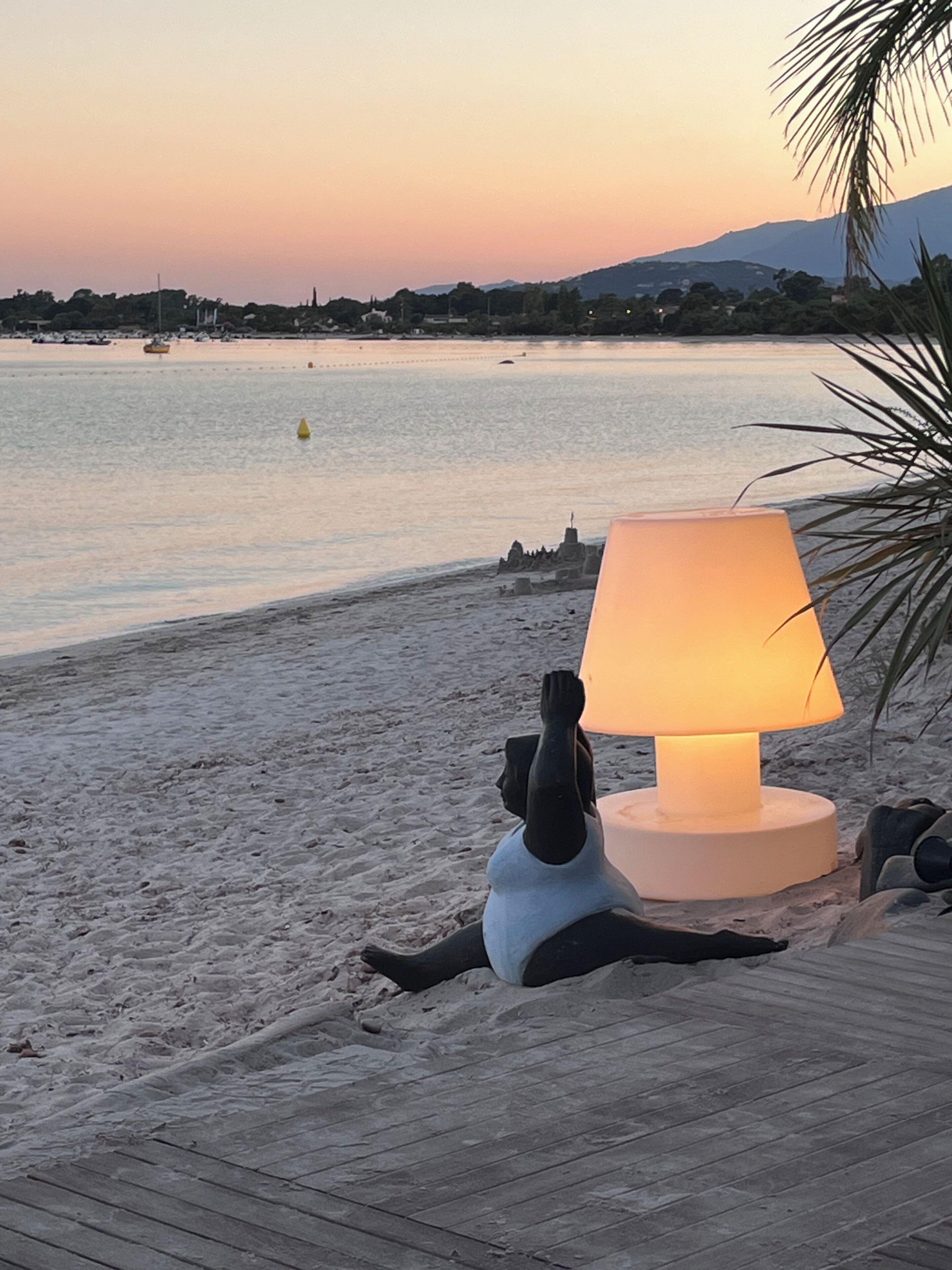 A person reclining on a wooden deck with a beach view, a lamp, and a palm tree in the foreground.