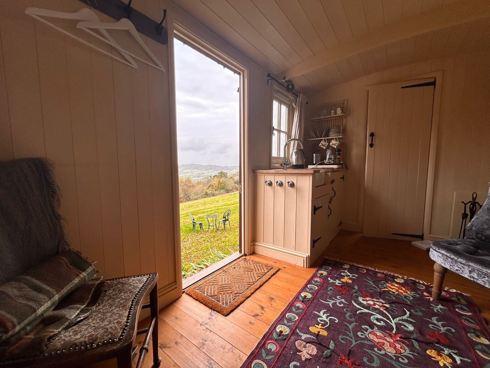 A cozy room with a wooden floor, a patterned rug, a chair, a sofa, and a view of a green landscape through a large window.