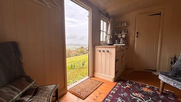 A cozy room with a wooden floor, a patterned rug, a chair, a sofa, and a view of a green landscape through a large window.
