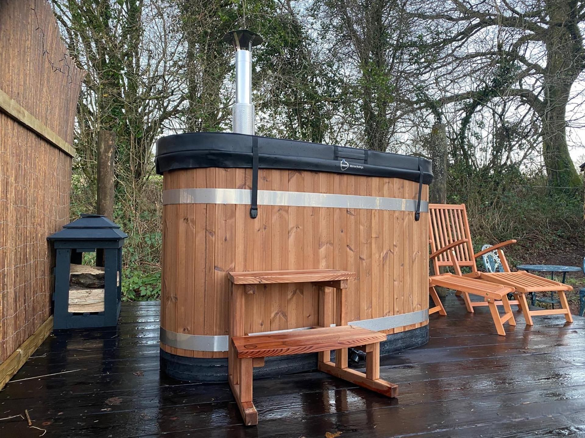 A wooden hot tub with a bench and a chair, surrounded by trees.