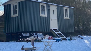 A small, single-story wooden structure with a white door and windows, a small porch with a table and chairs, and a wooden staircase leading up to the door.