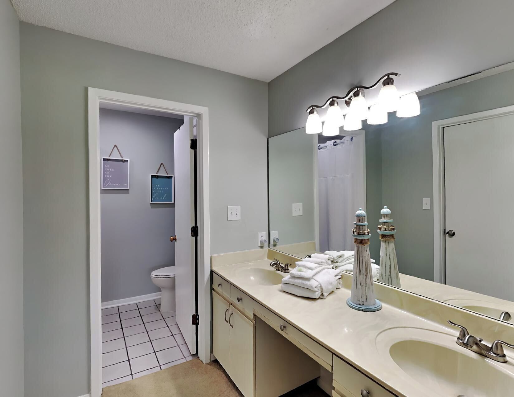 A bathroom with a large mirror, a sink, and a toilet. There are towels stacked on the sink and a framed quote on the wall.