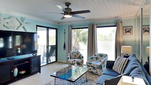 A living room with a TV, sofa, and chairs, a ceiling fan, and large windows. - Turtle Cay Golf Villa Edgewater Beach Resort (Panama City Beach)