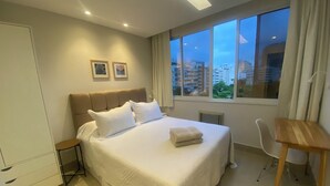 A hotel room with a large bed, a desk, and a view of the city at dusk. - Three Suites just a few steps from the beach (Ipanema Rio de Janeiro)