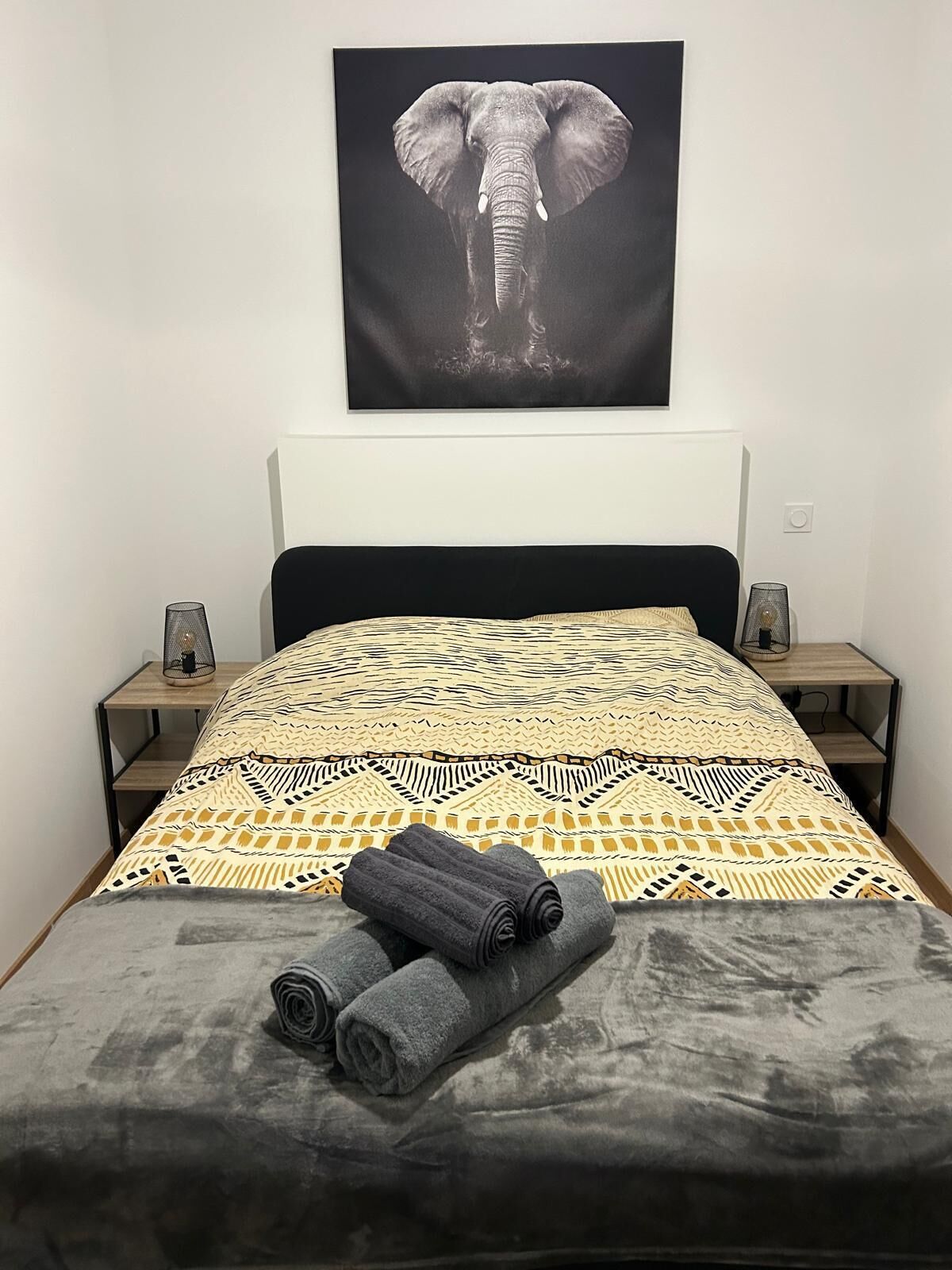 A bedroom with a patterned bedspread, a grey blanket, and rolled towels on the bed.