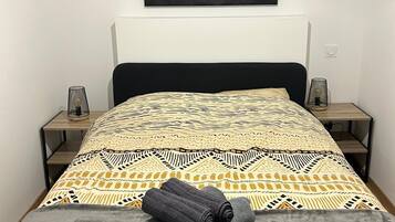 A bedroom with a patterned bedspread, a grey blanket, and rolled towels on the bed.