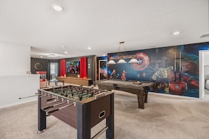 Quarto com mesa de pebolim, mesa de bilhar e um mural de super-heróis.