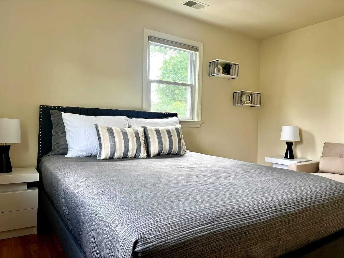 A neatly made bed with a grey and white striped pillow set, a bedside table with a lamp, and a window with blinds.