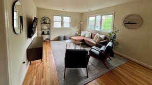 Ein geräumiges Wohnzimmer mit einer Couch, einem Couchtisch und einem Fernseher. Fenster sorgen für natürliches Licht und einen Ausblick nach draußen.