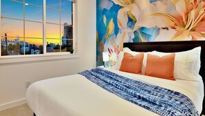 Ett hotellrum med ett stort fönster, en säng med vita och orange sängkläder och en väggmålning med blommotiv.
