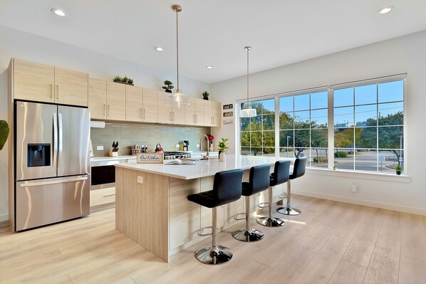 A modern kitchen with a stainless steel refrigerator, wooden cabinets, a large island with bar stools, and a view through large windows.